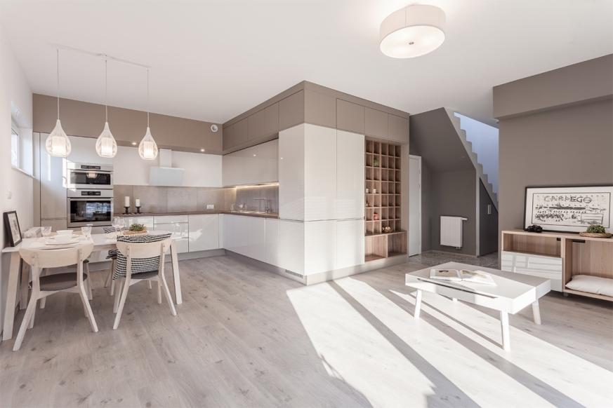 Nowoczesna, jasna kuchnia z białymi, lakierowanymi frontami szafek, drewnianą podłogą i stołem z nakryciem, oświetlona trzema wiszącymi lampami w kształcie kropli.