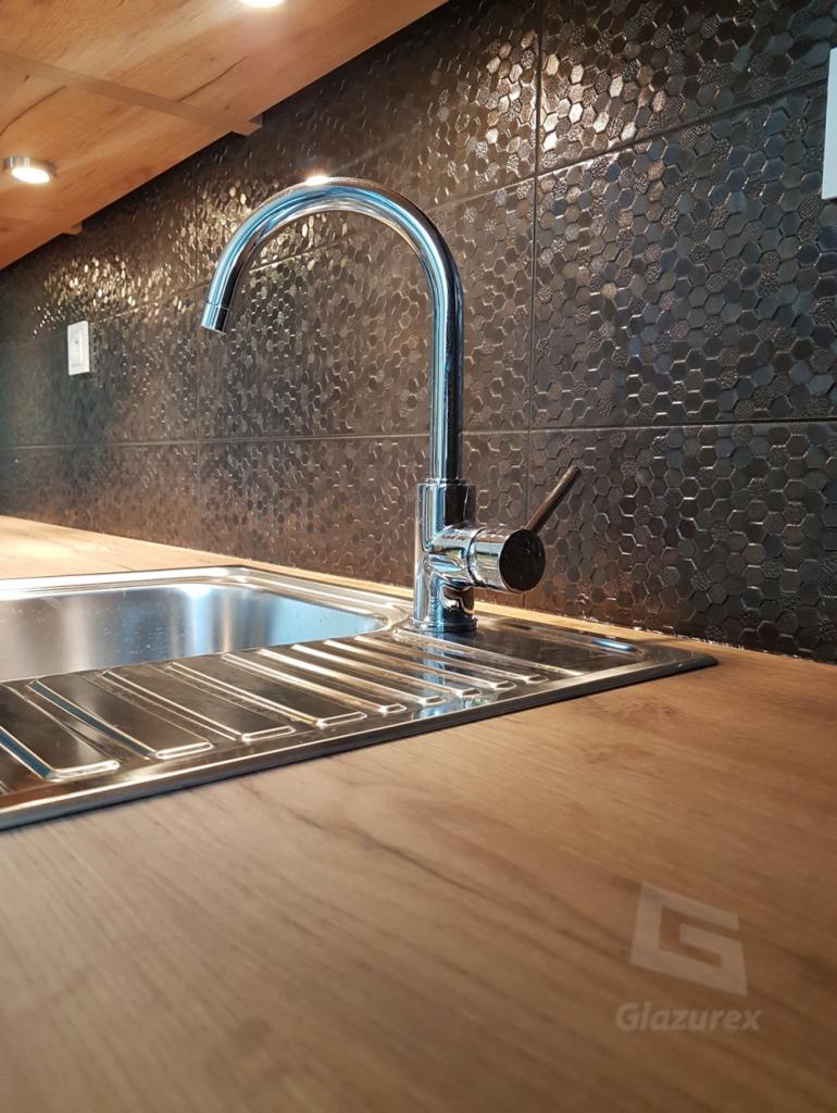 Bateria kuchenna ze stali nierdzewnej zamontowana w zlewozmywaku, na blacie z imitacji drewna, w tle ściana wyłożona czarnymi, heksagonalnymi płytkami ceramicznymi z fakturą.
