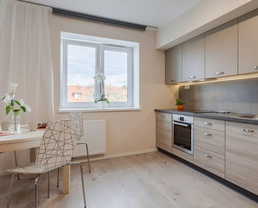 Jasna kuchnia z drewnopodobnymi frontami szafek, szarym blatem i oświetleniem LED pod szafkami wiszącymi, widok na okno z białymi firankami i kwiatami, stół z krzesłami w geometryczne wzory.