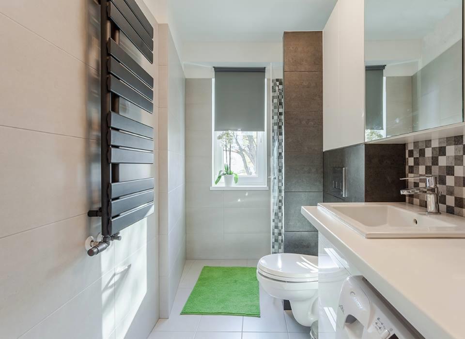 Jasna łazienka z grzejnikiem drabinkowym, mozaiką w strefie prysznica i zielonym dywanikiem przed oknem z roletą.