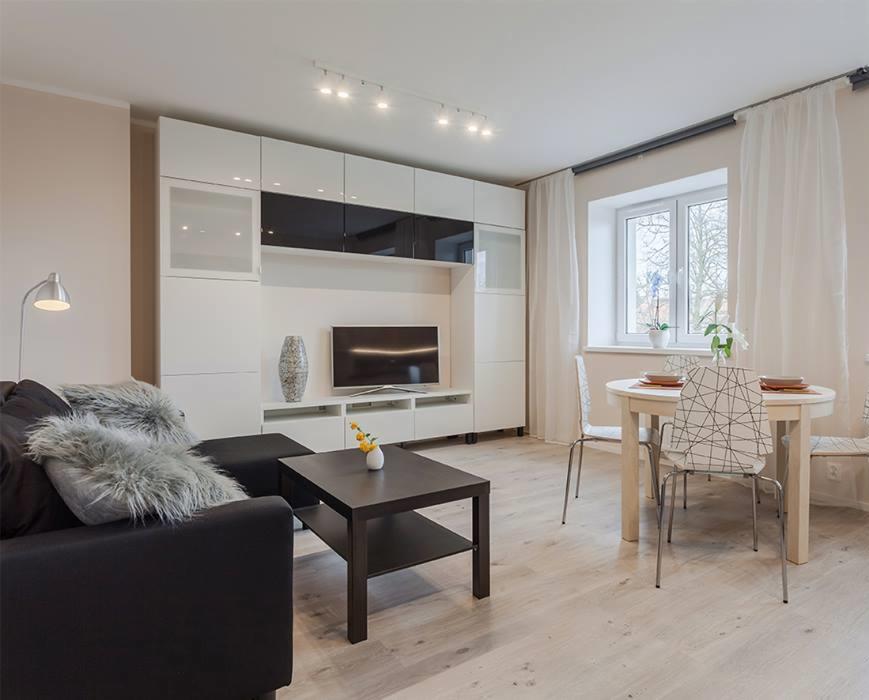 Jasne wnętrze salonu z białymi meblami modułowymi, czarną sofą z futrzanymi poduszkami, okrągłym stołem i panelami podłogowymi w odcieniach szarości.