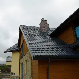 Nowoczesny dom z ciemnoszarą dachówką, drewnianą elewacją i ceglanym kominem, widok z dołu na tle pochmurnego nieba.
