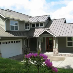 Dom jednorodzinny z szarym dachem z blachy trapezowej, widoczne rynny i elementy wykończeniowe, kwiaty rododendronu na pierwszym planie.