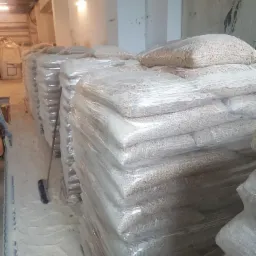 Magazyn z rzędami work&oacute;w wypełnionych pelletem, owiniętych folią, z widoczną miotłą opartą o jeden z work&oacute;w.