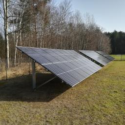 ArkonTech Paulina Kędzia - Rząd paneli fotowoltaicznych na łące, widok z boku, w tle las mieszany bez liści i ogrodzenie z siatki.