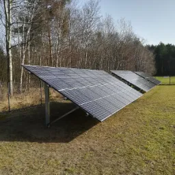 Rząd paneli fotowoltaicznych na łące, widok z boku, w tle las mieszany bez liści i ogrodzenie z siatki.