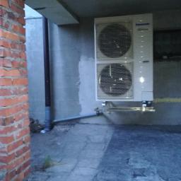 ArkonTech Paulina Kędzia - Biała, dwuczęściowa jednostka zewnętrzna klimatyzatora Panasonic zamontowana na ścianie szarego budynku, widoczna rura odprowadzająca skropliny, obok ceglany filar.