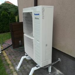 ArkonTech Paulina Kędzia - Zewnętrzna jednostka klimatyzacji Panasonic Inverter, zamontowana na białym stelażu z regulowanymi nóżkami, umieszczona na kostce brukowej obok ściany budynku z widoczną rurą odprowadzającą.