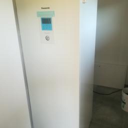 ArkonTech Paulina Kędzia - Biała jednostka wewnętrzna pompy ciepła Panasonic Aquarea z niebieskim wyświetlaczem, zamontowana w pomieszczeniu z szarą podłogą i białymi ścianami, widoczny fragment instalacji elektrycznej po...