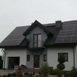 ArkonTech Paulina Kędzia - Dom jednorodzinny z zamontowanymi panelami fotowoltaicznymi na dachu, widoczny numer domu 65B i mężczyzna w tle na podjeździe.