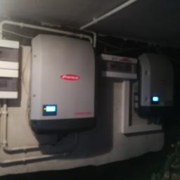 ArkonTech Paulina Kędzia - Dwa białe inwertery marki Fronius zamontowane na szarej ścianie z widocznymi rurami instalacyjnymi i skrzynkami elektrycznymi. Jeden inwerter jest większy i ma czerwone logo, drugi mniejszy. Oba...