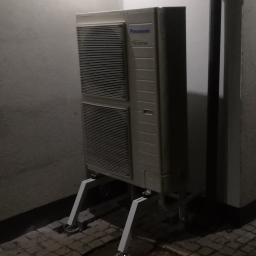 ArkonTech Paulina Kędzia - Zewnętrzna jednostka klimatyzacji Panasonic Inverter, zamontowana na specjalnym stelażu z kółkami, ustawiona przy ścianie budynku z kostki brukowej.