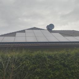 ArkonTech Paulina Kędzia - Panele fotowoltaiczne na brązowym dachu domu jednorodzinnego, widoczne z poziomu gruntu na tle gęstego żywopłotu i pochmurnego nieba.