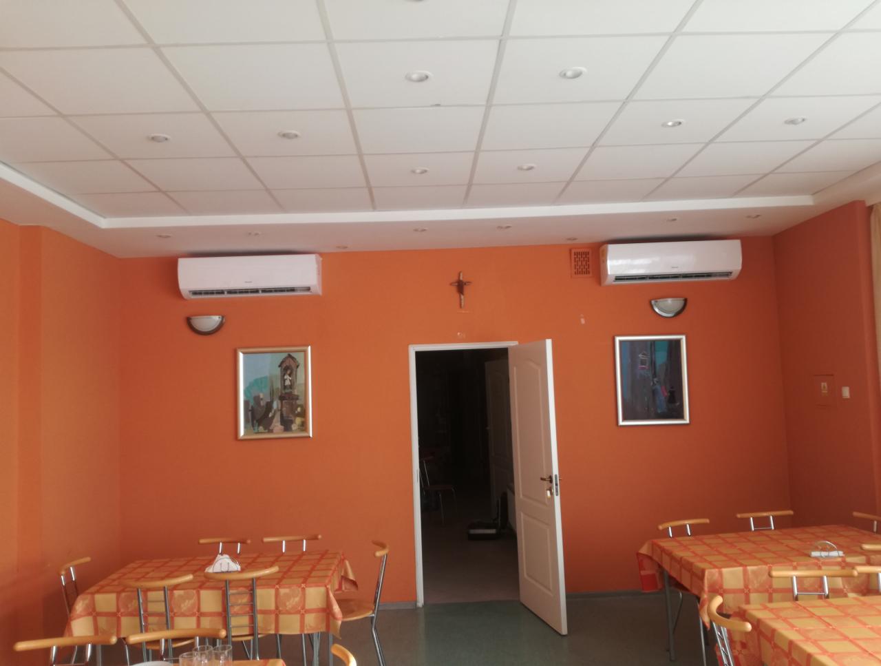 Pomarańczowa sala jadalna z dwoma zainstalowanymi klimatyzatorami ściennymi, oświetleniem sufitowym i obrazami na ścianach.