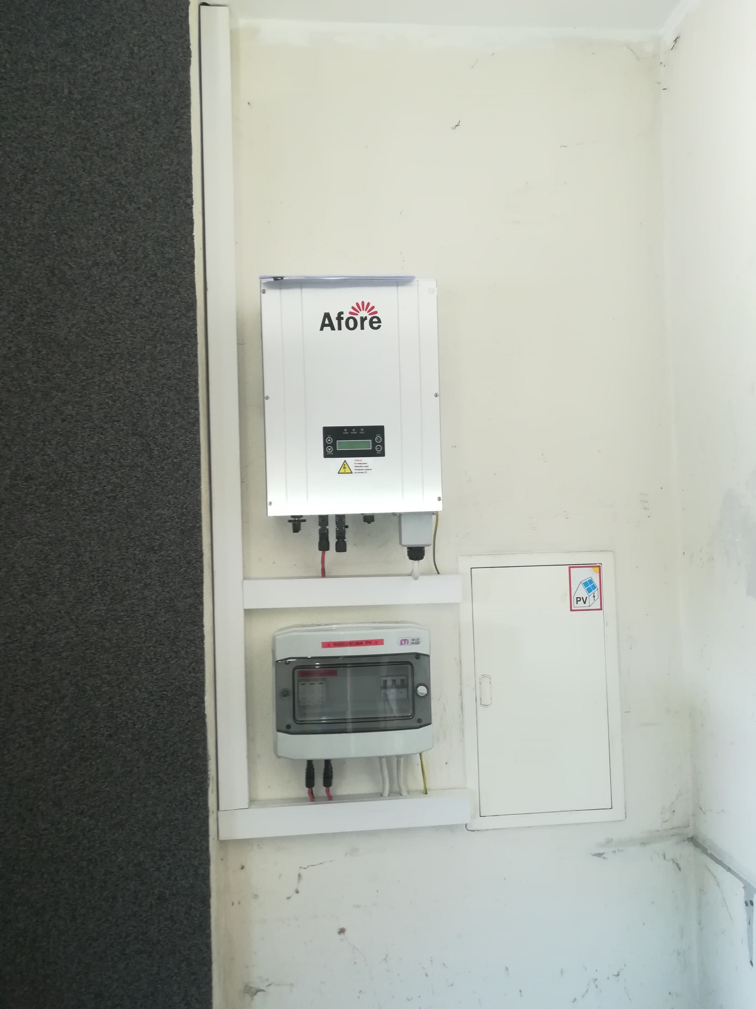 Zamontowany inwerter Afore i skrzynka bezpiecznikowa na ścianie w pomieszczeniu, widoczny także fragment ściany pokrytej szarą wykładziną.