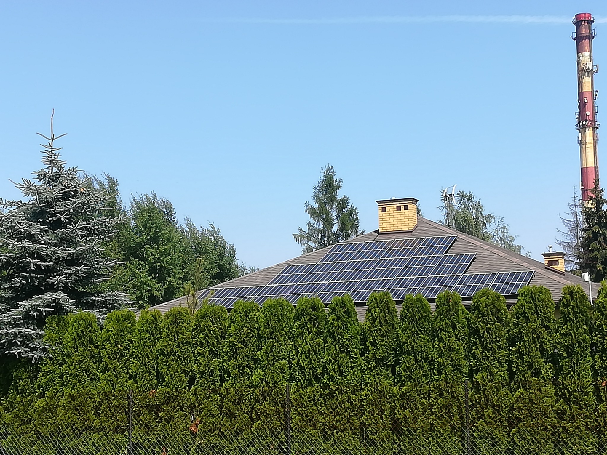 Dom jednorodzinny z brązowym dachem pokrytym panelami słonecznymi, widoczne drzewa iglaste i komin przemysłowy w tle.