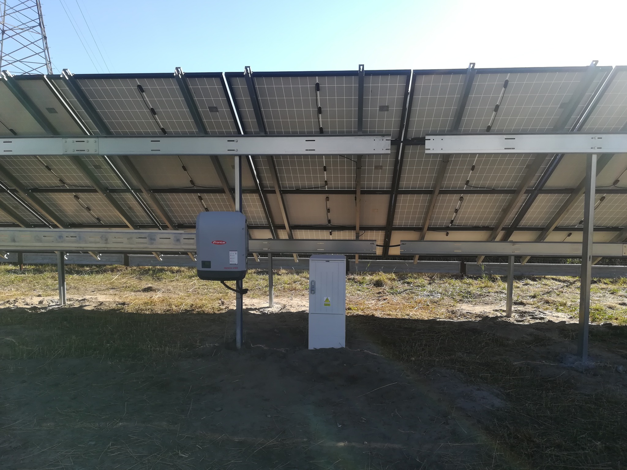 Widok od spodu na panele fotowoltaiczne zamontowane na metalowej konstrukcji, widoczny inwerter Fronius i skrzynka elektryczna z ostrzeżeniem o napięciu, w tle słup wysokiego napięcia.