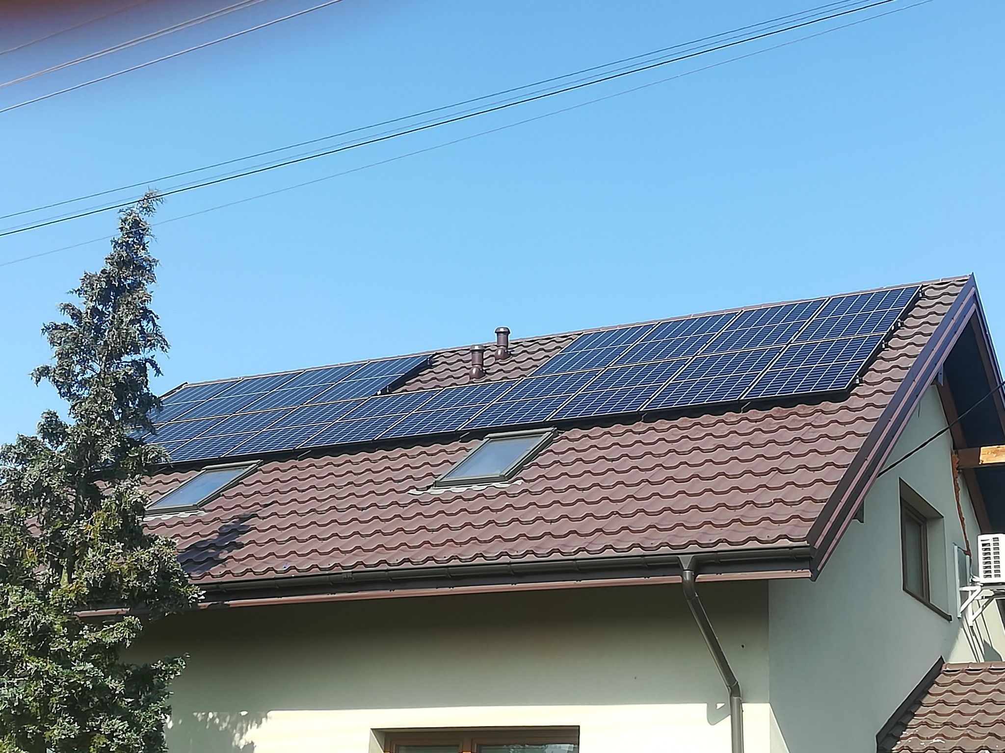 Dom z brązowym dachem, oknami dachowymi i panelami słonecznymi, z klimatyzatorem na ścianie bocznej.