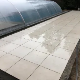 Wyłożony jasnymi, kwadratowymi płytkami taras przy basenie z zadaszeniem, widoczne odbicia drzew na mokrej powierzchni płytek, obrzeże tarasu w trakcie wykańczania.