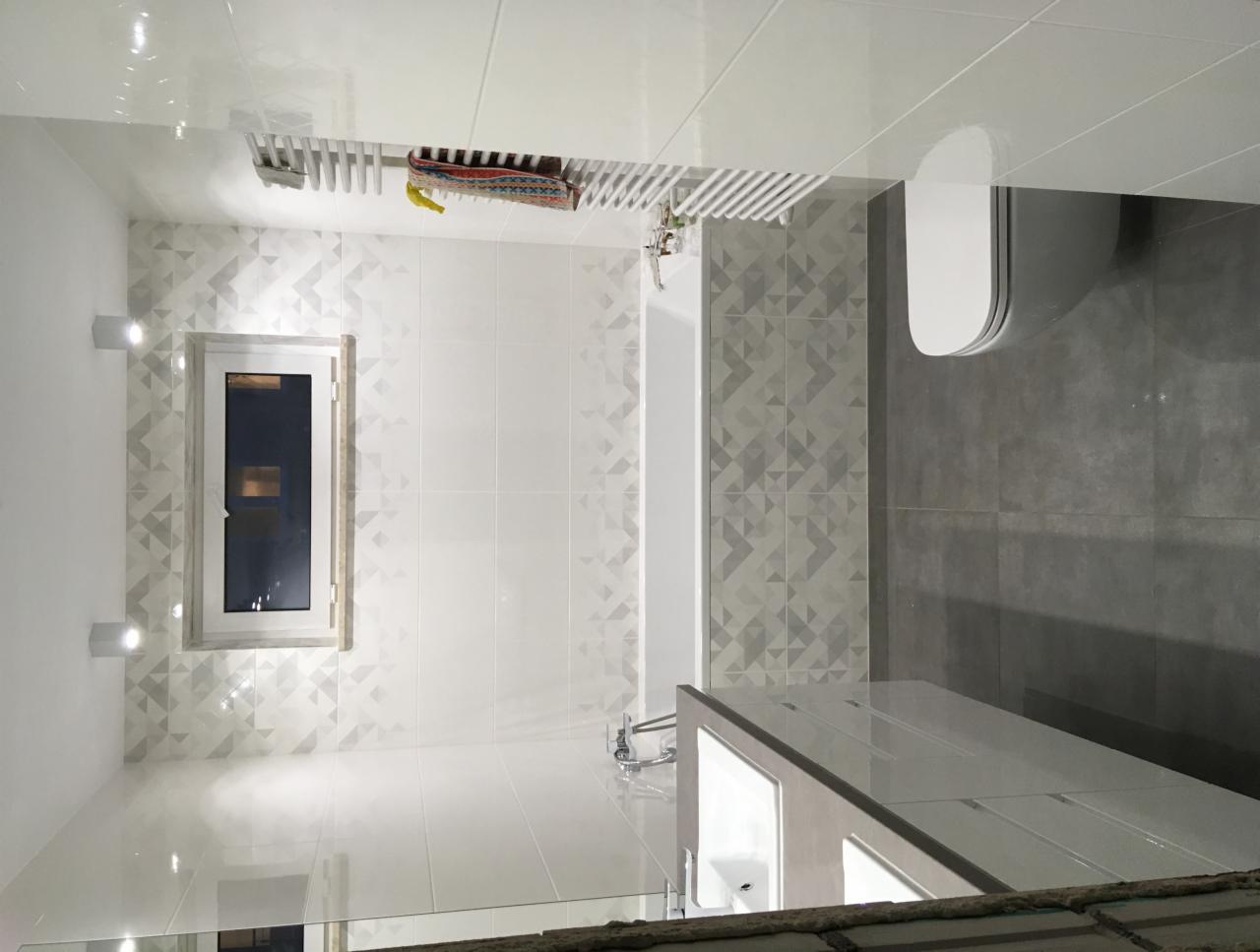 Nowoczesna łazienka z geometrycznym wzorem na białych płytkach, szara podłoga i biała armatura. Widoczny fragment umywalki z lustrem, toaleta, wanna, okno z oświetleniem punktowym i grzejnik...