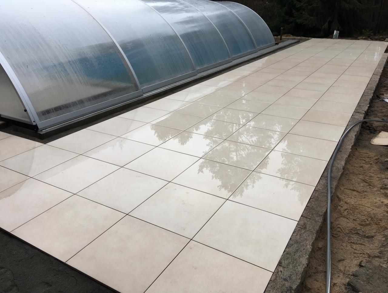 Wyłożony jasnymi, kwadratowymi płytkami taras przy basenie z zadaszeniem, widoczne odbicia drzew na mokrej powierzchni płytek, obrzeże tarasu w trakcie wykańczania.