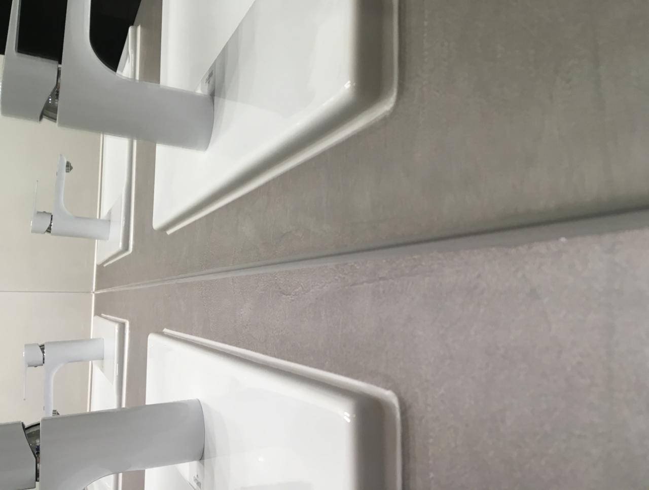 Rząd umywalek z białymi bateriami zamontowanych na szarej ścianie z płytek, widok z dołu pod kątem.