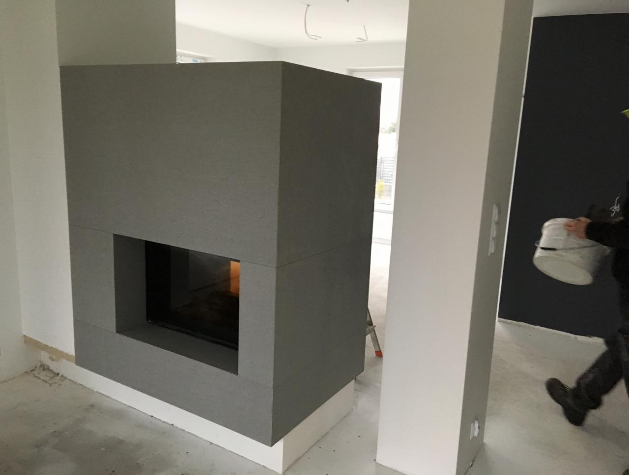 Nowoczesny, szary kominek z wkładem, w trakcie prac wykończeniowych, widoczny fragment pomalowanej na ciemny kolor ściany i osoba z wiadrem farby w tle.