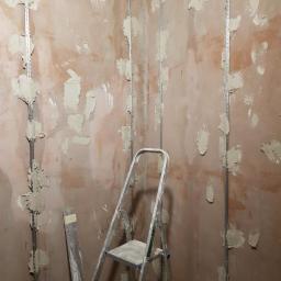 Budrom - Przygotowanie ściany pod montaż płyt karton gips: metalowe profile przytwierdzone do surowej ściany z widocznymi plamami zaprawy, drabina aluminiowa w rogu pomieszczenia.