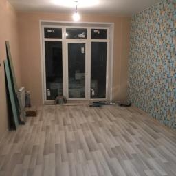 Budrom - Pokój po remoncie: panele podłogowe ułożone, jedna ściana z wzorzystą tapetą, pozostałe pomalowane na beżowo, przy ścianie oparte płyty karton gips, widoczne narzędzia i materiały budowlane.