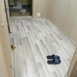 Budrom - Jasnoszare panele podłogowe ułożone w wąskim przedpokoju, widoczne niebieskie klapki i narzędzia przy ścianie, ściany beżowe.