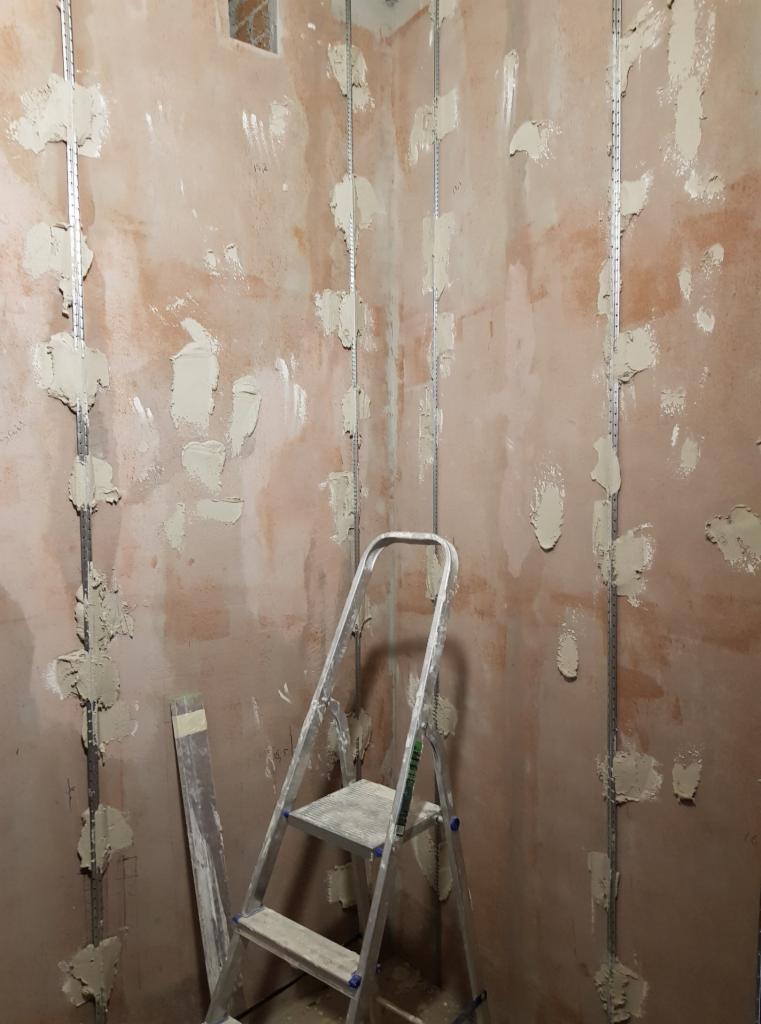 Przygotowanie ściany pod montaż płyt karton gips: metalowe profile przytwierdzone do surowej ściany z widocznymi plamami zaprawy, drabina aluminiowa w rogu pomieszczenia.