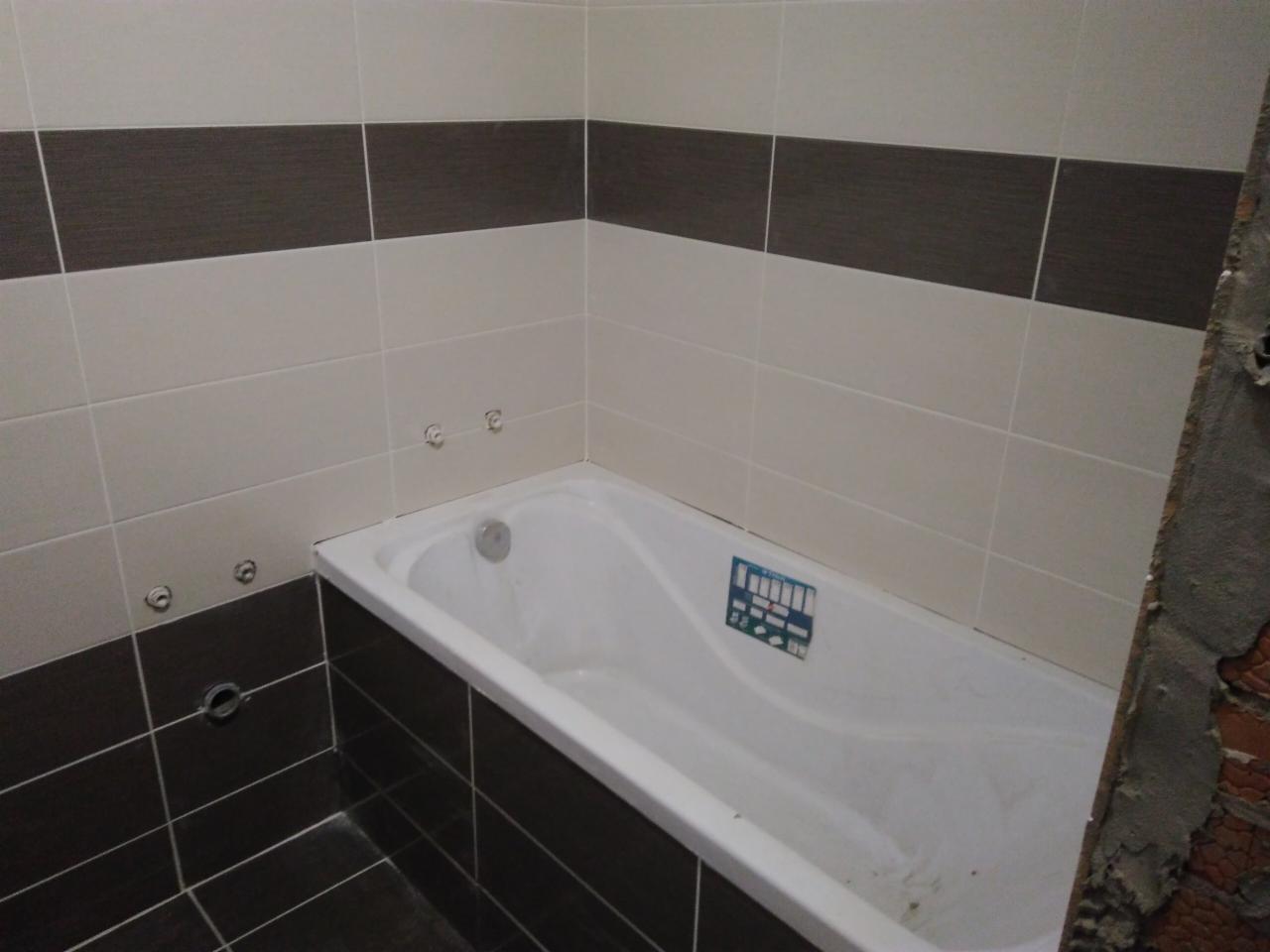 Fragment łazienki w trakcie wykańczania: ułożone dwukolorowe płytki (beżowe i brązowe) na ścianach i podłodze, biała wanna z niebieską naklejką, widoczne przygotowane punkty instalacyjne, surowy...