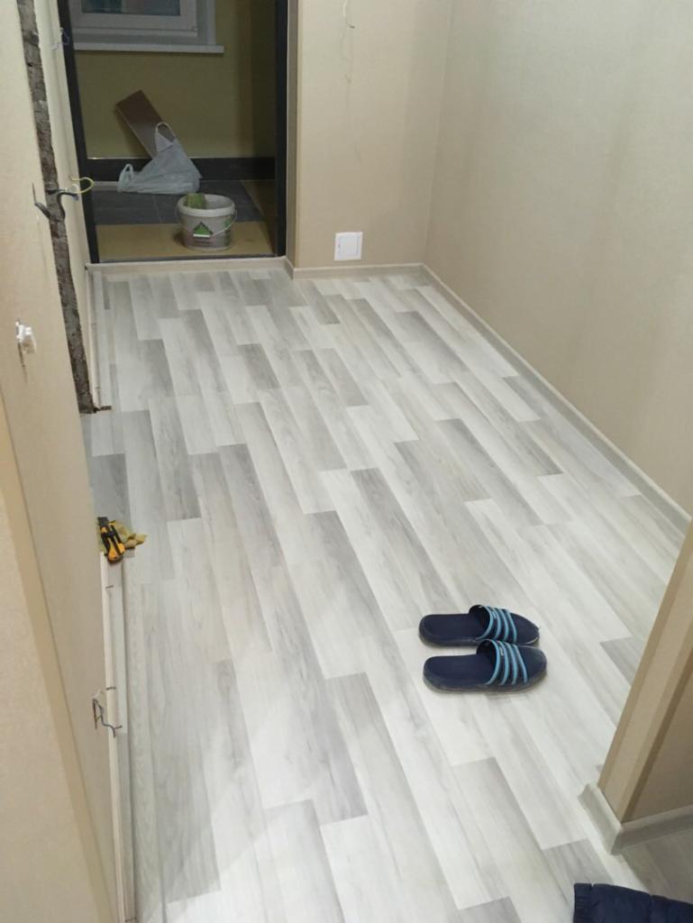 Jasnoszare panele podłogowe ułożone w wąskim przedpokoju, widoczne niebieskie klapki i narzędzia przy ścianie, ściany beżowe.