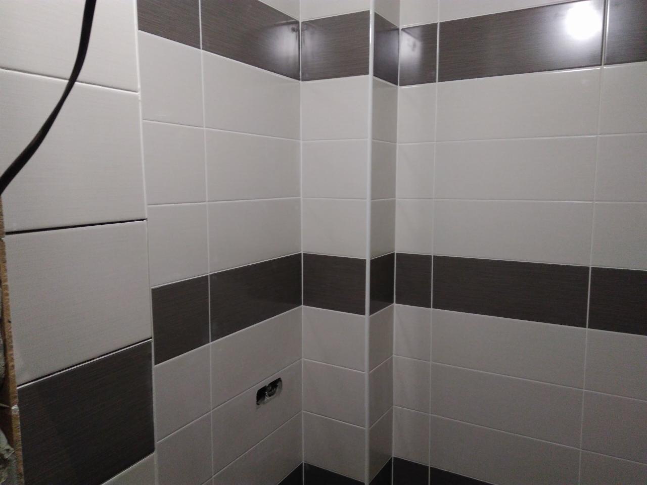 Wykończona łazienka z białymi i brązowymi płytkami ułożonymi w poziome pasy, widoczny fragment instalacji elektrycznej w ścianie.