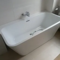 Biała, prostokątna wanna z chromowaną baterią, zamontowana w wykafelkowanej łazience z beżowymi płytkami podłogowymi i białymi płytkami ściennymi.