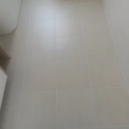 Jasne, kwadratowe płytki podłogowe w łazience, widok z góry, fuga w kolorze zbliżonym do płytek, fragment wanny i szafki w tle.