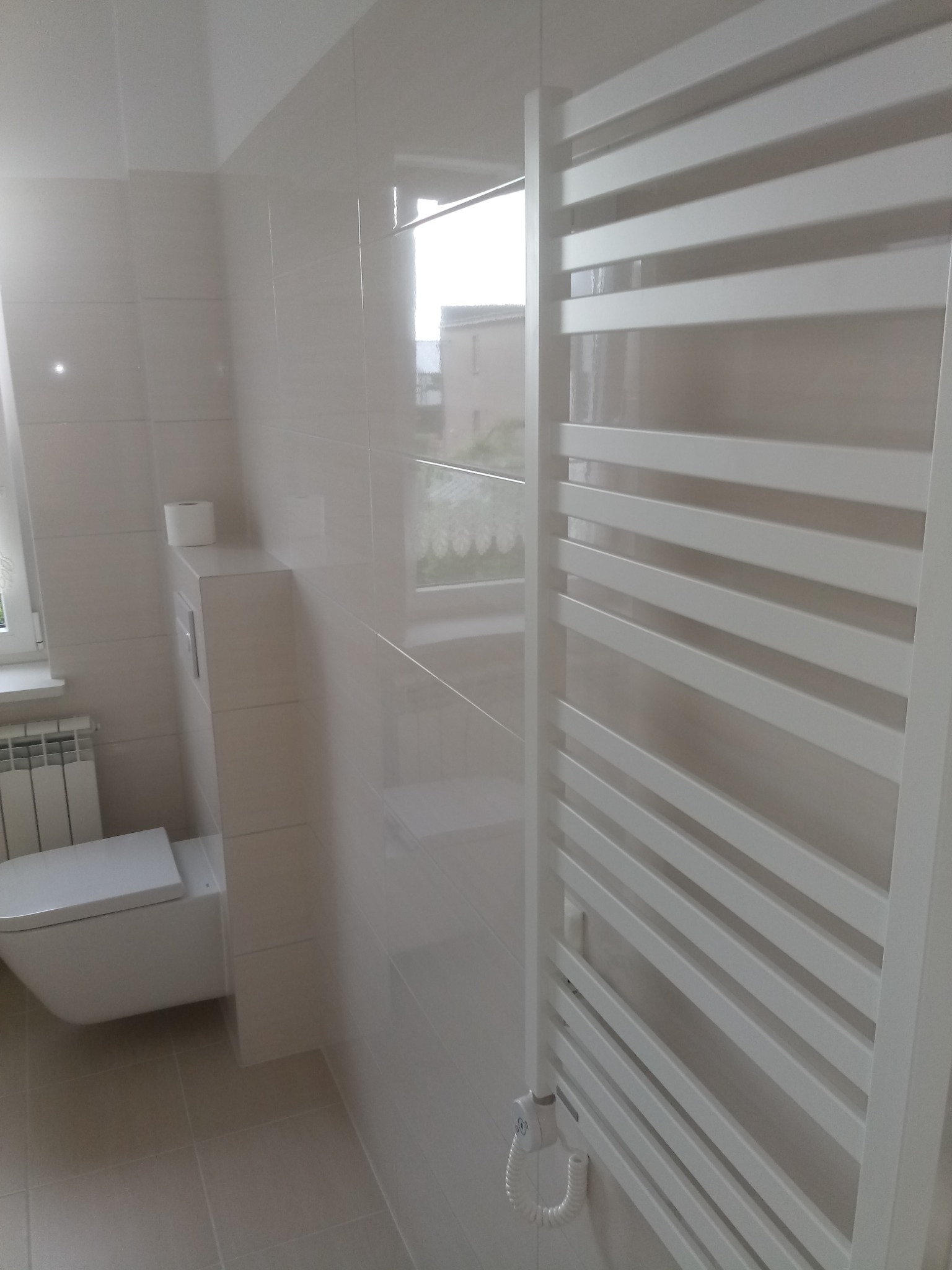 Biała łazienka z błyszczącymi, beżowymi płytkami, wiszącą toaletą, grzejnikiem drabinkowym i oknem z widokiem na budynek.