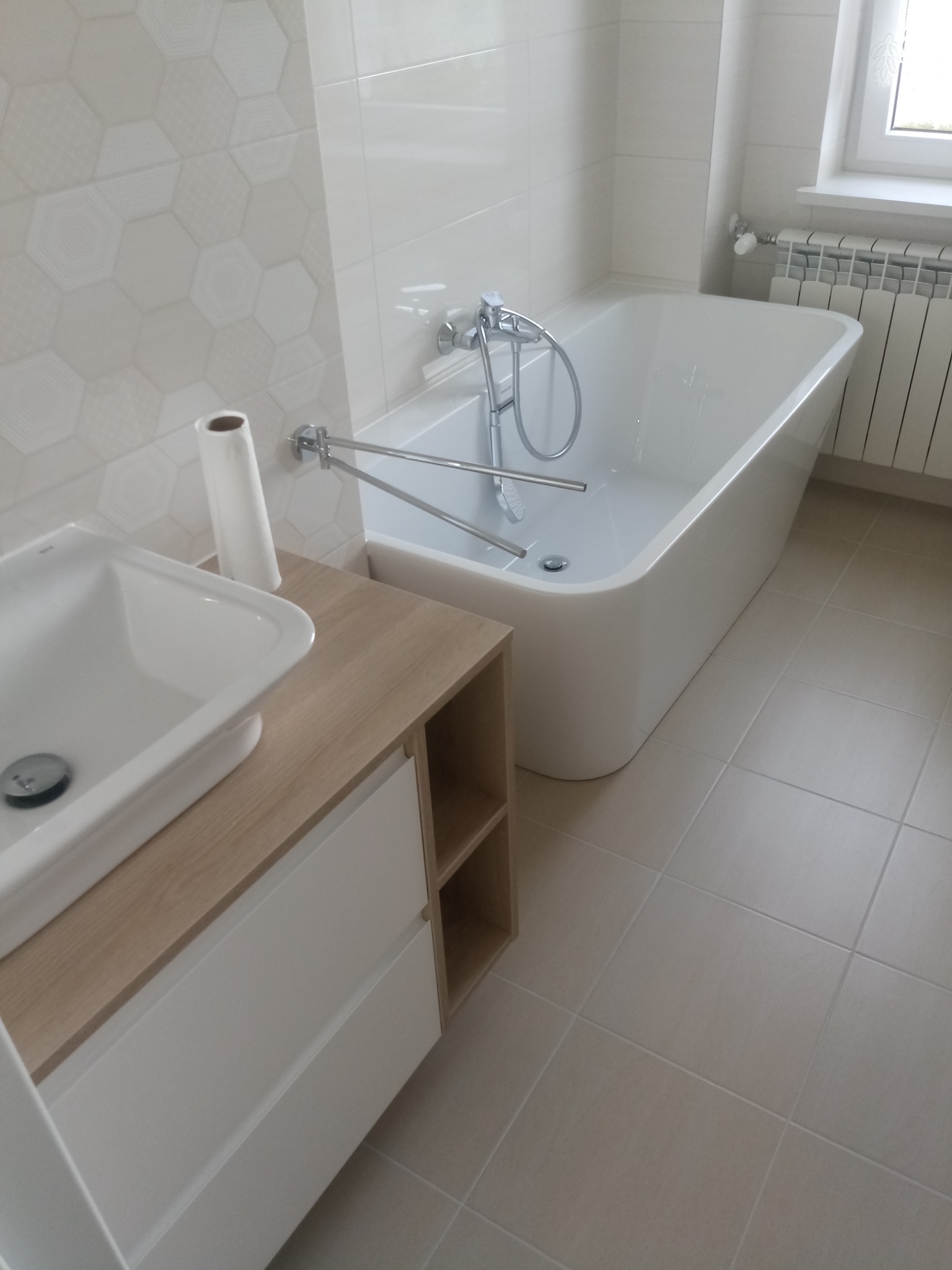 Nowoczesna łazienka z białą wanną, umywalką nablatową na szafce z drewnianym blatem i heksagonalnymi płytkami na ścianie, uzupełniona o grzejnik drabinkowy.
