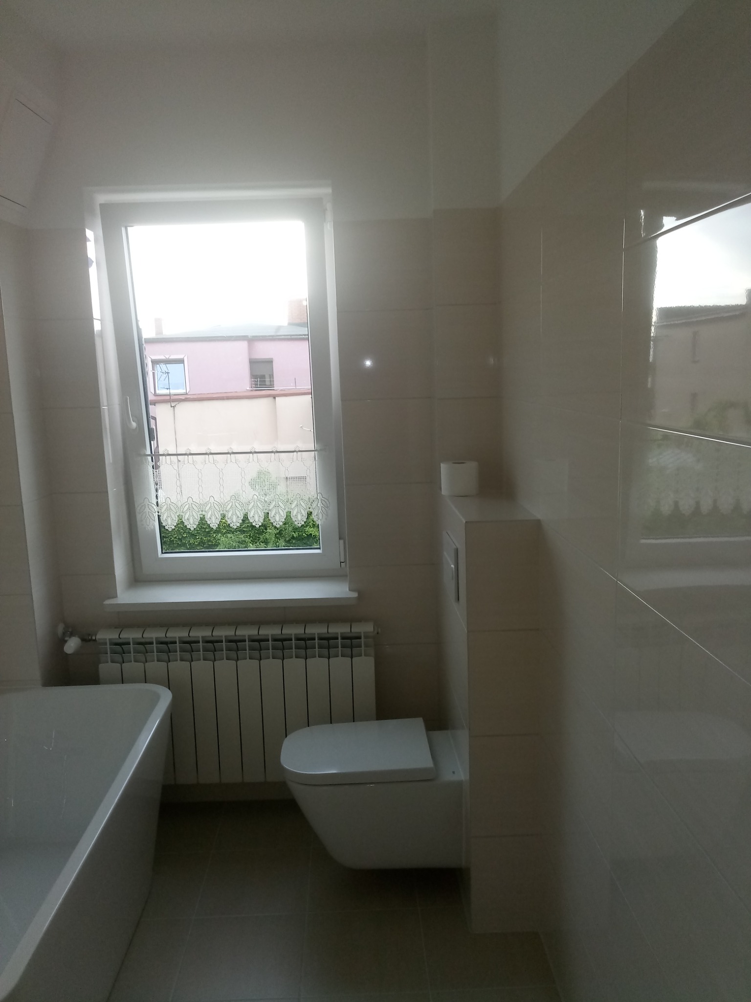 Jasna łazienka z beżowymi płytkami, białą wanną, toaletą i grzejnikiem pod oknem z firanką; widok na różowy budynek za oknem.