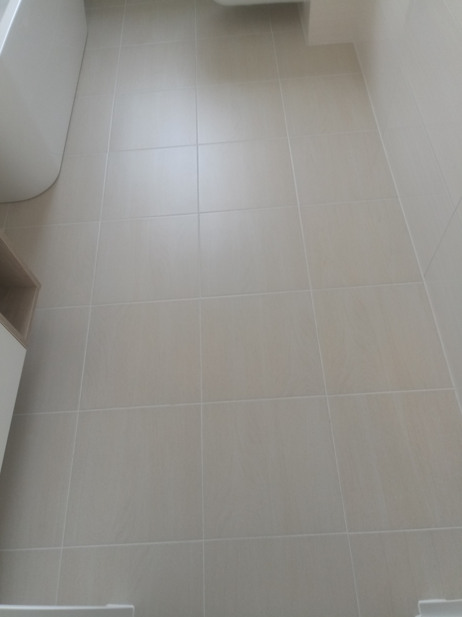 Jasne, kwadratowe płytki podłogowe w łazience, widok z góry, fuga w kolorze zbliżonym do płytek, fragment wanny i szafki w tle.