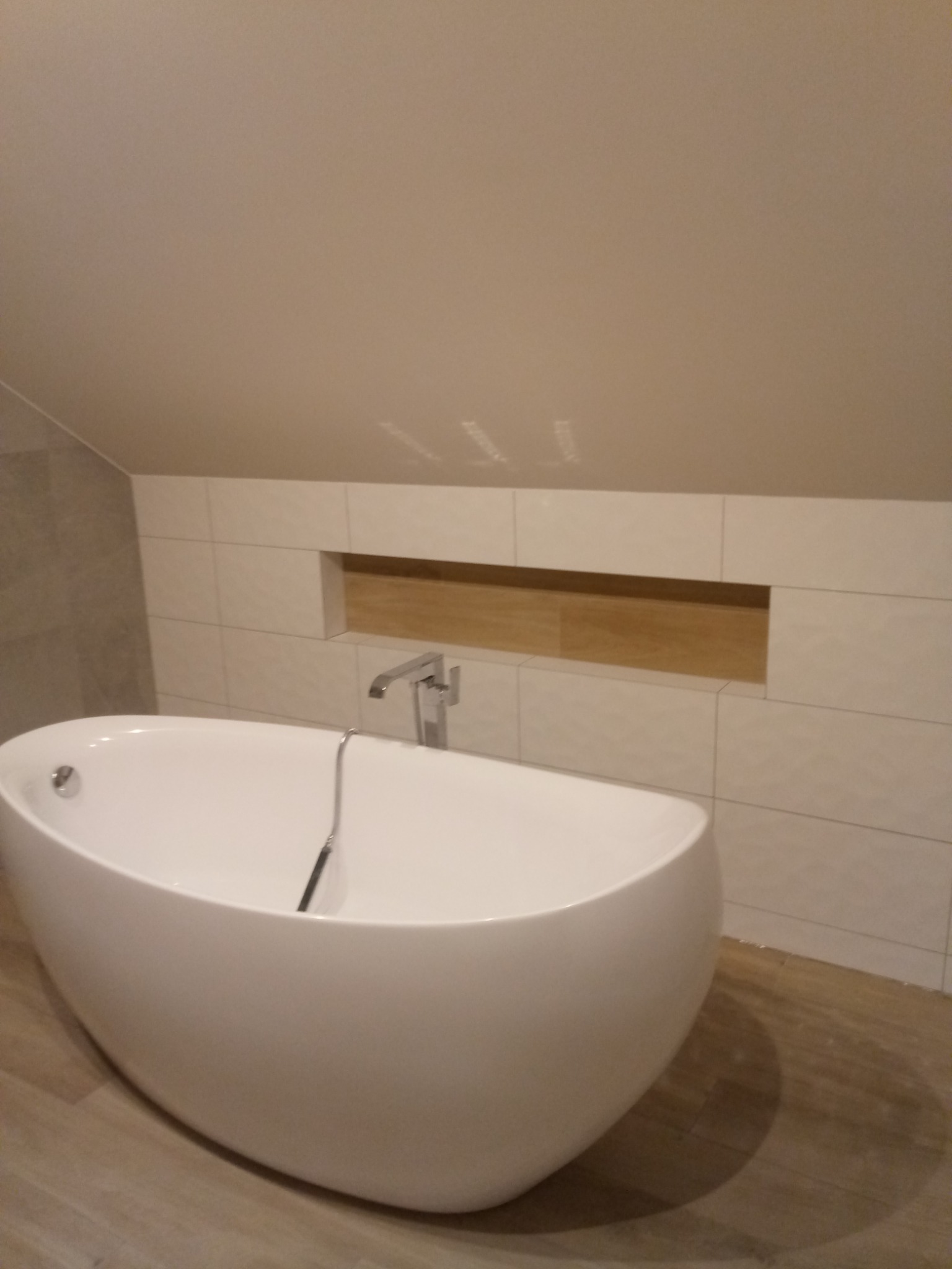 Biała, wolnostojąca wanna owalna z chromowaną baterią w łazience na poddaszu, wykończonej płytkami w odcieniach beżu, z wnęką w ścianie wyłożoną drewnem.