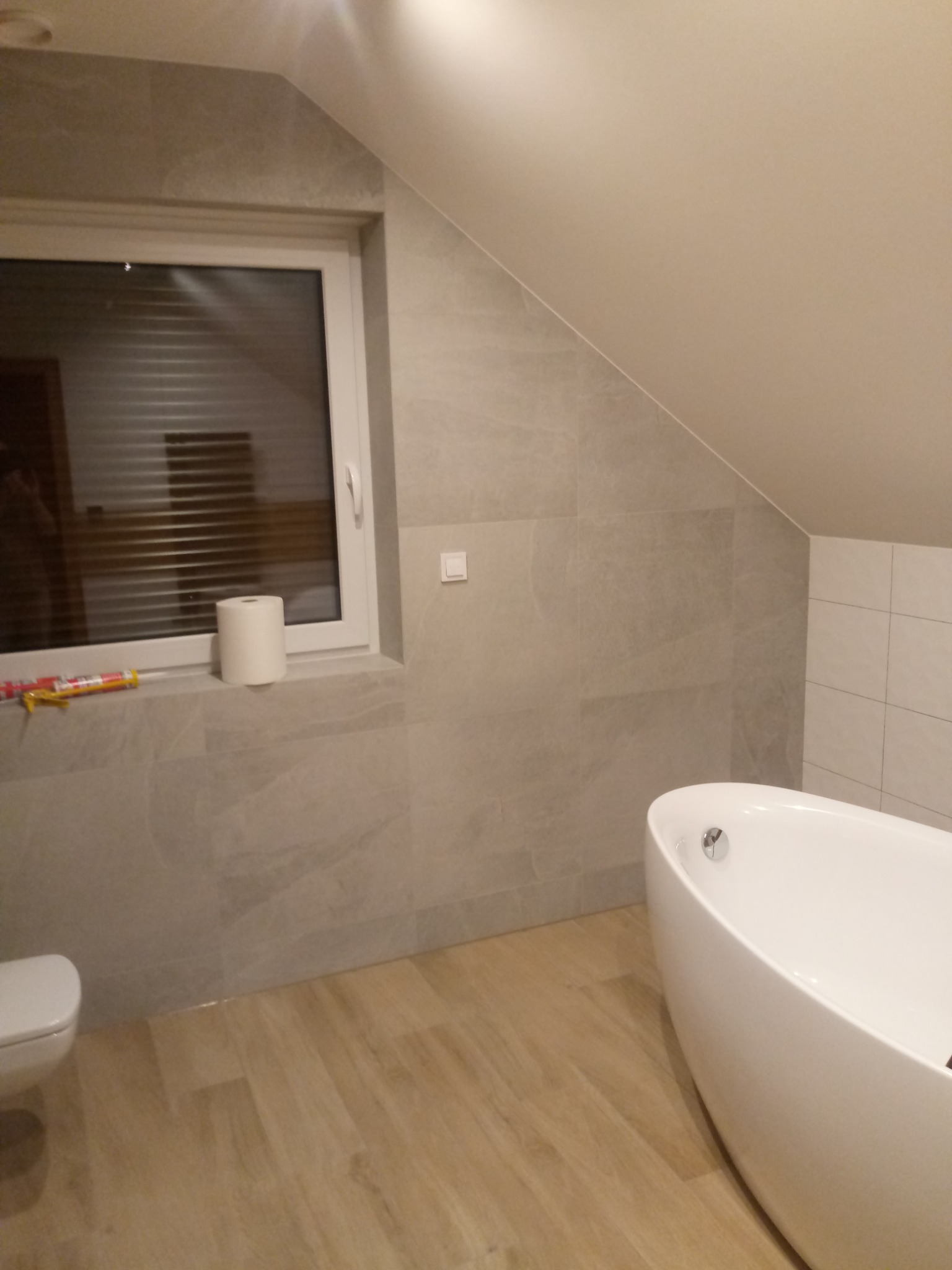 Jasna łazienka na poddaszu z wanną wolnostojącą, wykończona płytkami imitującymi kamień i drewnopodobną podłogą, z widocznym silikonem i papierem toaletowym na parapecie okna.