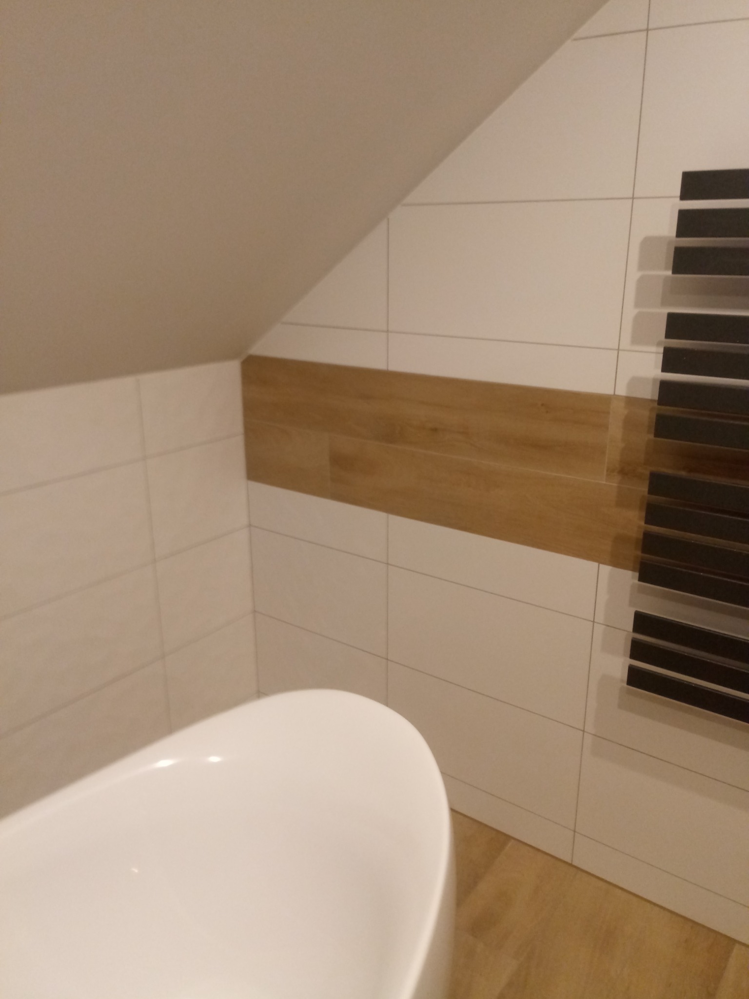 Biała wanna wolnostojąca w łazience na poddaszu, z białymi płytkami i poziomym pasem płytek imitujących drewno, obok grzejnik drabinkowy z czarnymi poprzeczkami.