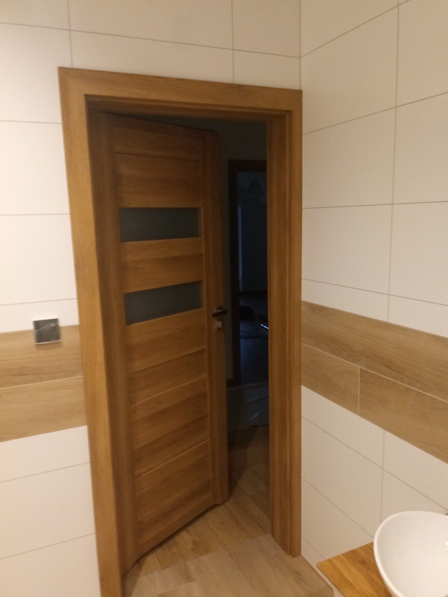 Otwarta drewniana ościeżnica z przeszklonymi panelami w łazience z białymi i beżowymi płytkami, widok na sąsiednie pomieszczenie