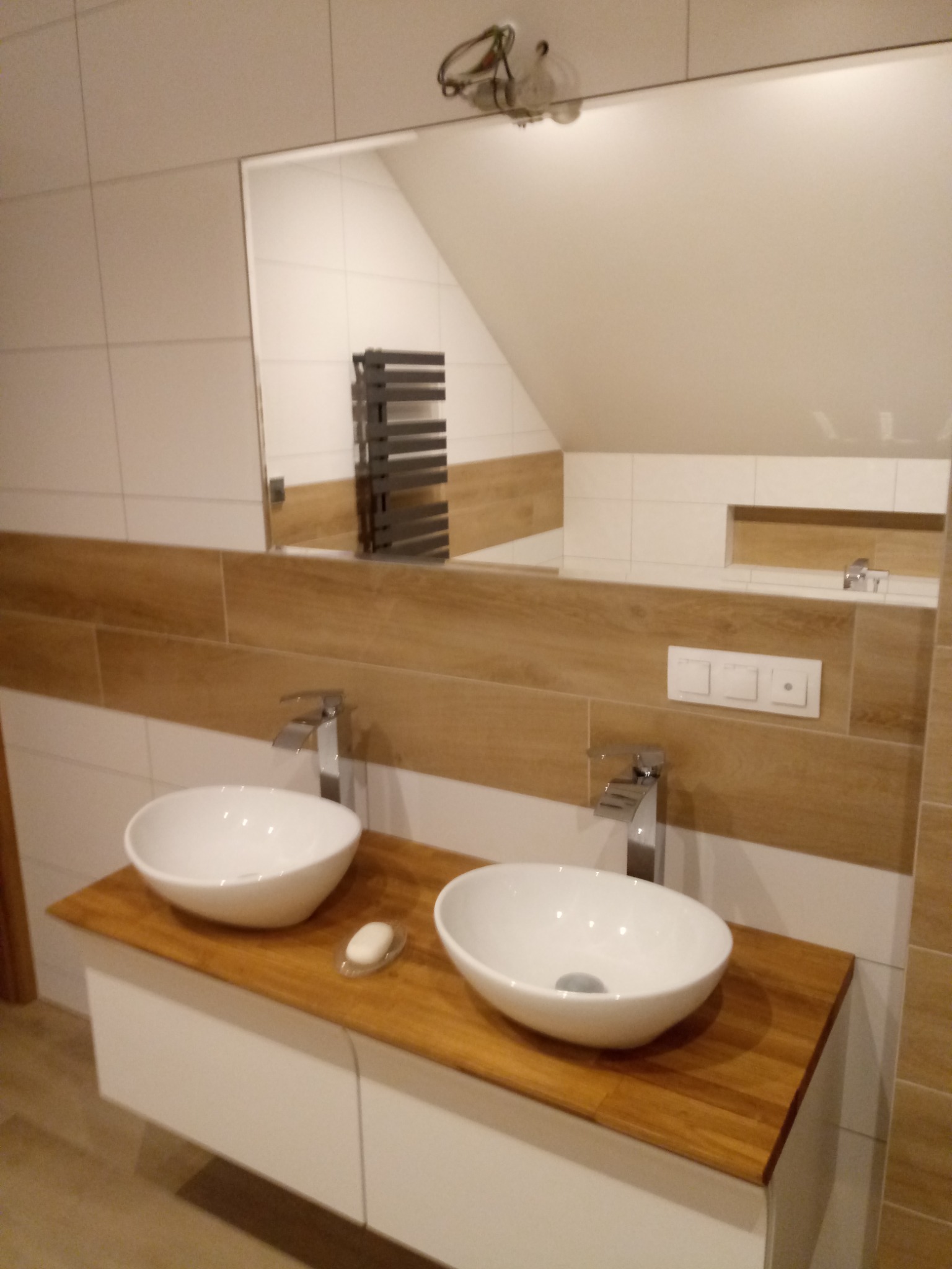 Nowoczesna łazienka z dwiema białymi umywalkami nablatowymi na drewnianym blacie, widoczne płytki imitujące drewno na ścianie, duże lustro odbijające grzejnik drabinkowy.