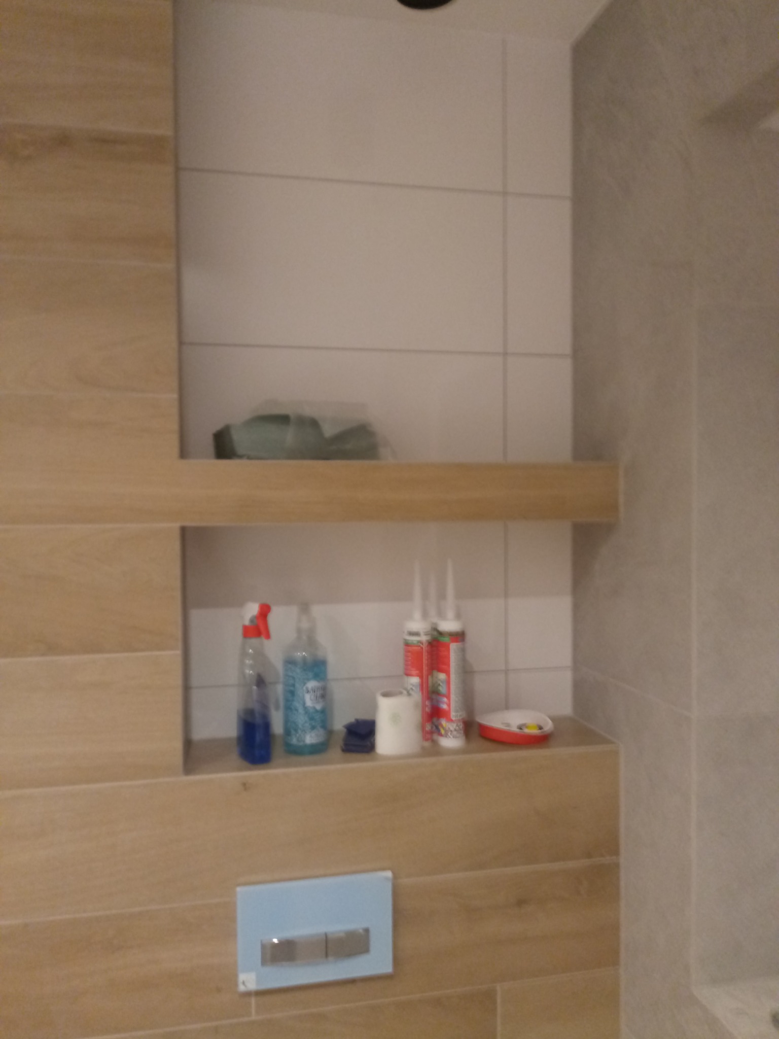 Półka z drewnopodobnych płytek w łazience z artykułami higienicznymi i materiałami budowlanymi, widok na przycisk spłuczki wbudowany w ścianę.