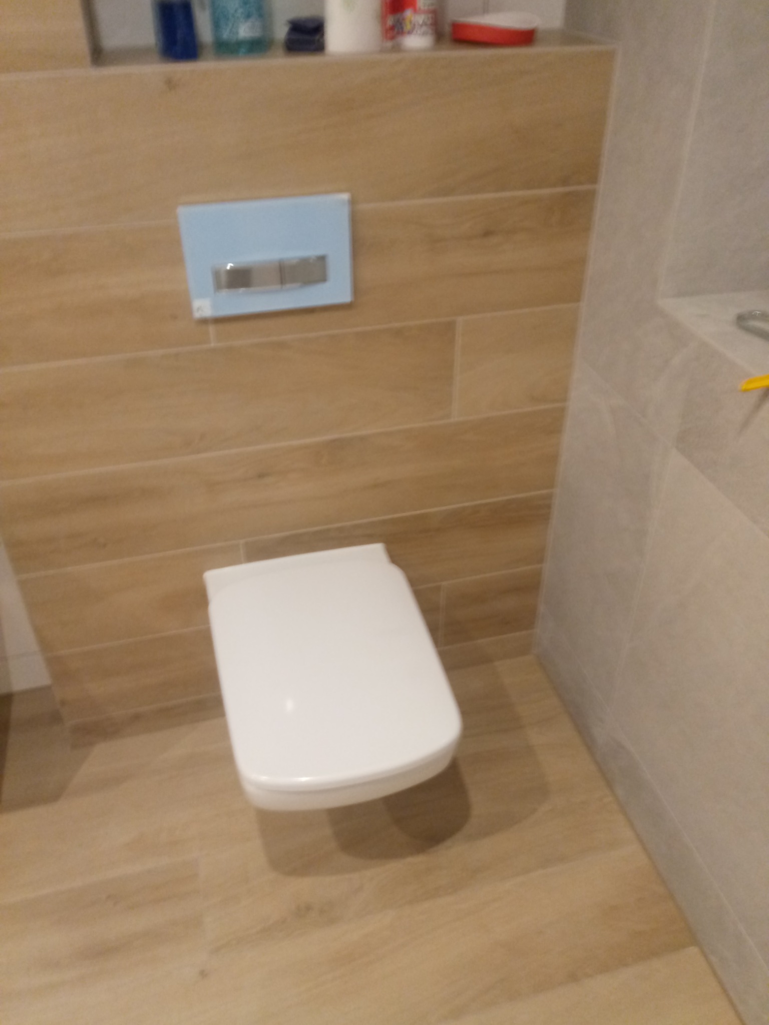 Podwieszana toaleta z deską wolnoopadającą, zamontowana na ścianie wyłożonej płytkami imitującymi drewno, z niebieskim panelem spłukującym.