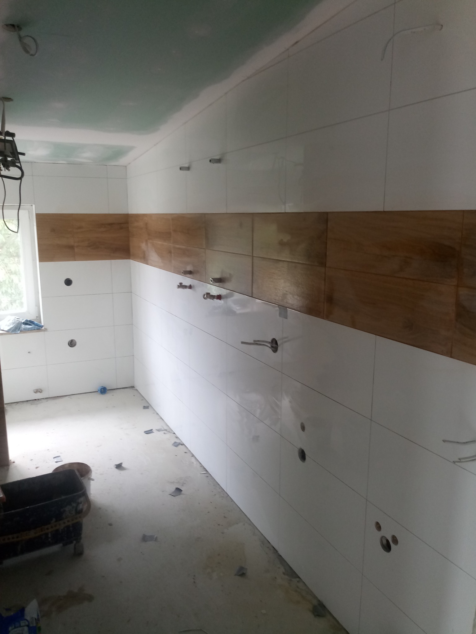 Wnętrze w trakcie wykańczania z częściowo położonymi białymi i drewnopodobnymi płytkami ściennymi, widoczne otwory na instalacje, surowy sufit i podłoga, oraz narzędzia pracy w rogu pomieszczenia.