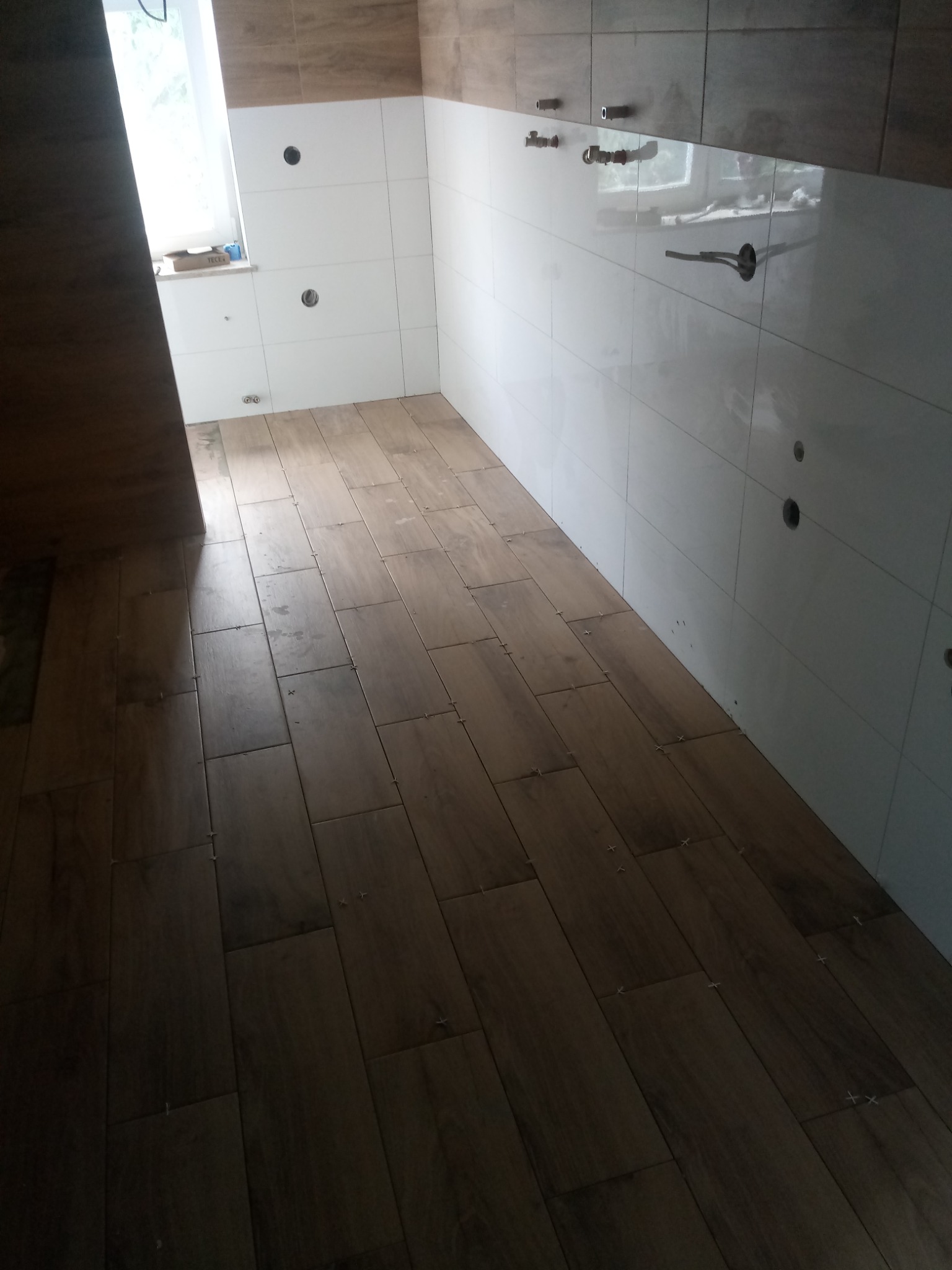 Układanie płytek podłogowych imitujących drewno w nowo wykończonej kuchni, widoczne białe płytki ścienne z przygotowanymi otworami na instalacje.