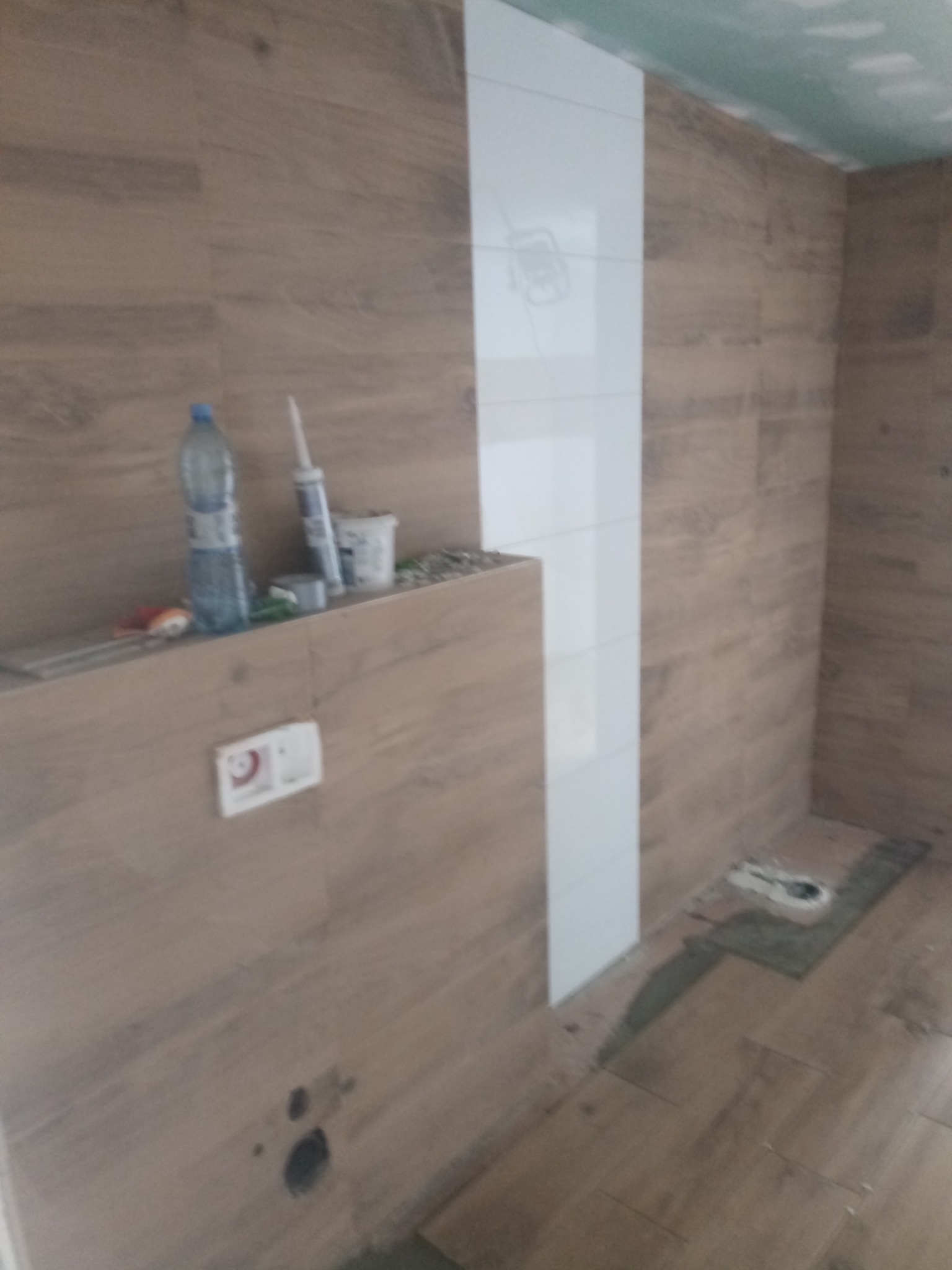 Montaż płytek ściennych imitujących drewno z pionowym pasem białych płytek w łazience w trakcie remontu, widoczne materiały budowlane i niedokończone prace.